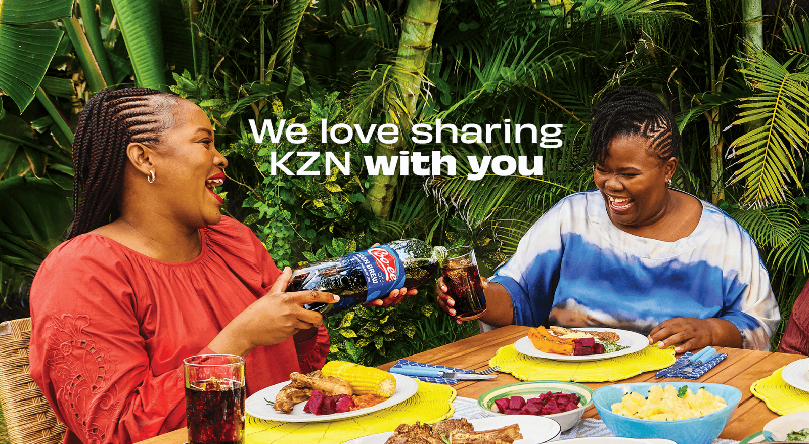 Two happy women sharing a meal and pouring Coo-ee soda outdoors, celebrating friendship and the joyful spirit of KZN together.